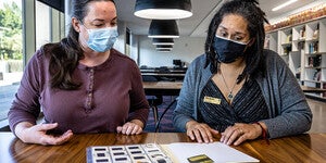 Two women wearing masks sitting at a table talking and looking at photographic slides