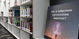 Library bookshelves filled with books behind a large poster with text "Why is Indigenous representation important?"