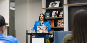 Woman in a blue jacket, standing at a podium, talking into a microphone in a library