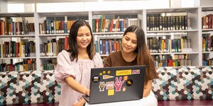 Two people sitting in the library looking at a laptop together with shelves of books behind them