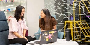 Two students sitting around a table looking at a laptop with the Makerspace 3D printers in the background