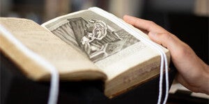 A person rests their hand on a book that is sitting on a book cradle, strings of page weights are on each side.