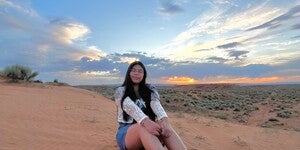 Photo of Myacedes Miller sitting on a hill composed of tan sand with footprints around her. She is sitting in front of a sunset wearing white shoes and jean shorts. The sky is blue with clouds blotting an orange sun low on the horizon.