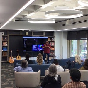 Delbert Anderson speaking in front of a crowd of people in the Labriola Center, room 204. Anderson is wearing a red shirt and holding a mic with his trumpet behind him.