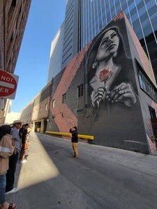 Photo of Breeze speaking in front of the second mural in downtown Phoenix. This mural is a black and white portrait of a young girl from the community holding a red rose looking East. The black and white and gray portrait contains spirals beneath, which weave throughout the mural and can be seen in the triangular border on either side of the girl.