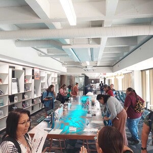 Pictured are participants surrounding the Labriola Center's Storytelling Table looking at Indigenous childrens books offered at the Office of Indian Education Symposium. The light from the table emits a turquoise glow.