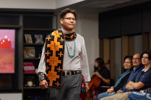 Model on a runway wearing a gray outfit with an orange and black geometric scarf, turquoise necklace, and metallic belt in front of a seated audience.