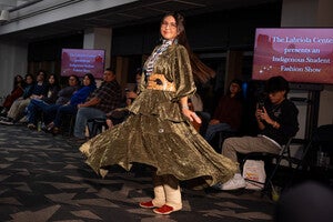 Model in an olive-green dress walks on a runway during a fashion show with seated audience.