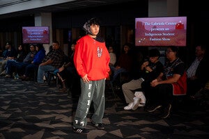 Model on a runway in red hoodie and gray green pants with seated audience and screens in background.