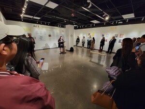 Large group of people facing a podium with speaker in a large open room with archive materials post on the walls.