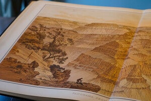 An oblique angled photograph of Panorama of Point Sublime Looking West. Prominent ridges, cliffs, and slopes of the Grand Canyon are variably orange and tan. Sitting on the ledge of the overlook, An artist sits sketching while a surveyor glances from behind. Agave plants and large oak trees are visible on the left side of the cliff's edge. 