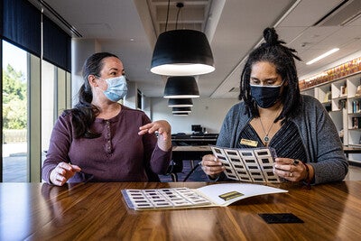 Two women wearing masks sitting at a table talking and looking at photographic slides