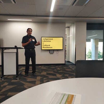 Pictured is Eric Hardy (Diné) presenting in room 236 for the Office of Indigenous Education Symposium. The slide behind him says "Indigenous Libraries = Cultural Resilience"