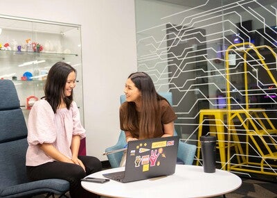 Two students sitting around a table looking at a laptop with the Makerspace 3D printers in the background