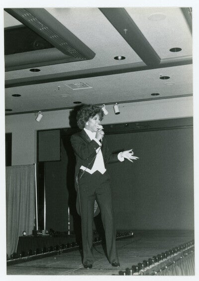 A gorgeous performer wearing a tailsuit and glamorous makeup sings to the crowd (1982)